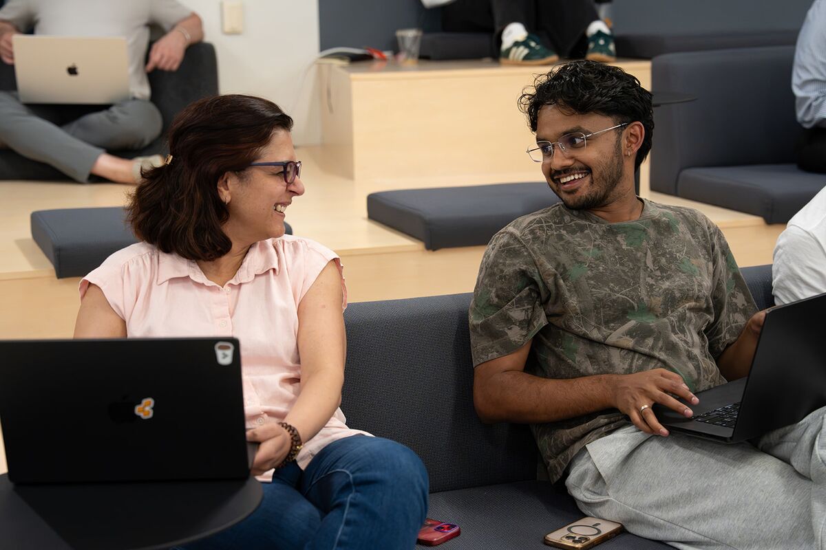 Two teammates laughing together while working on laptops in a lounge