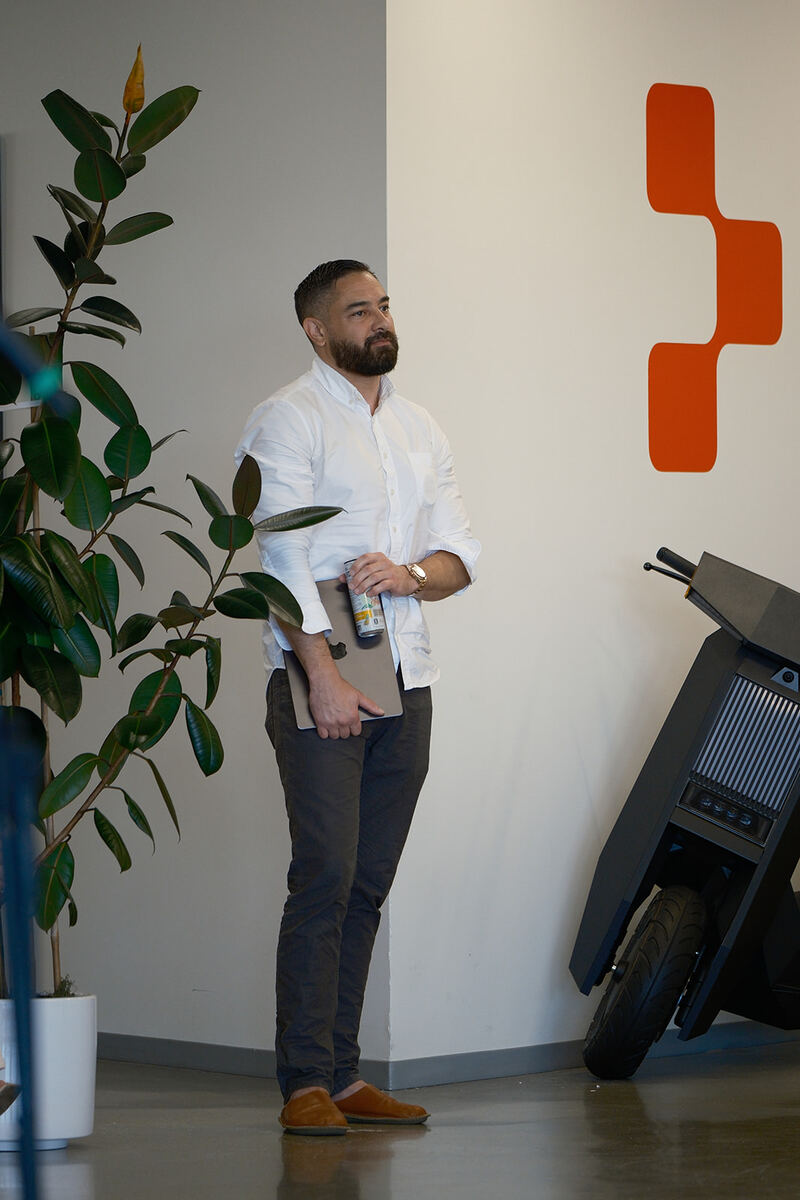 A teammate standing beside a plant and the orange Replit logo on an office wall